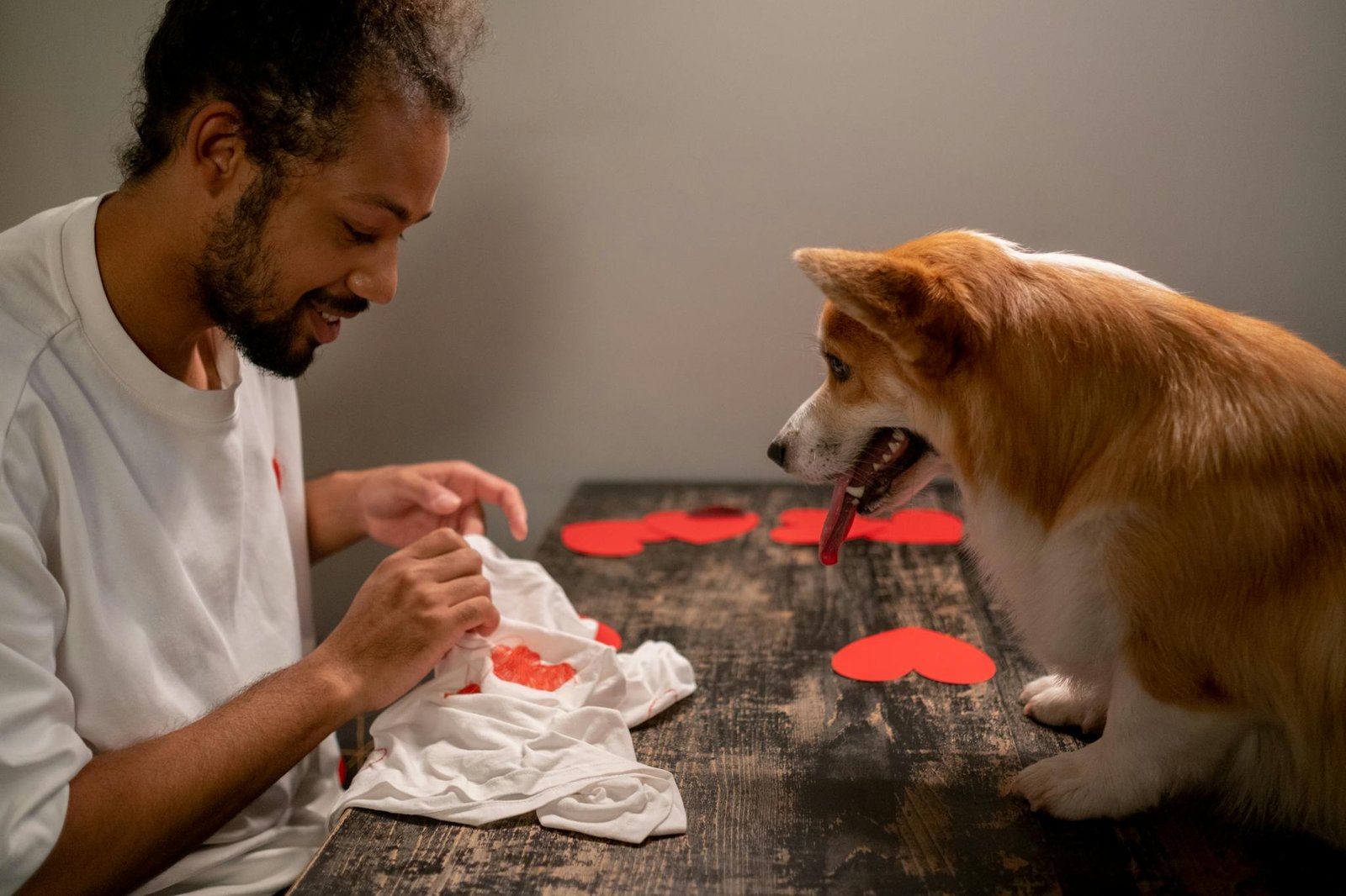 A man crafts heart decorations on a table indoors with his attentive dog nearby.