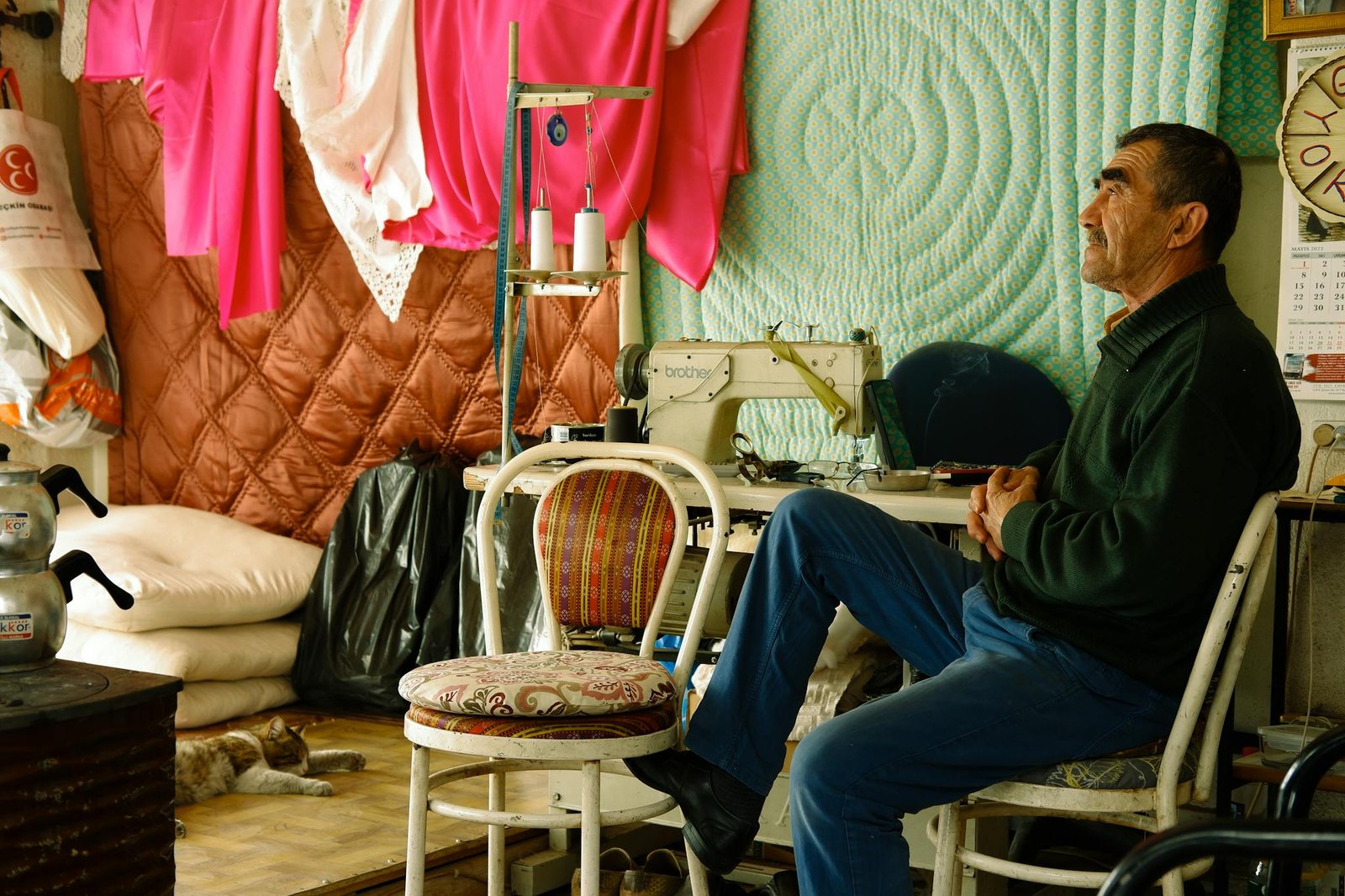 Elderly tailor sitting in his colorful sewing workshop with a cat and sewing machine.